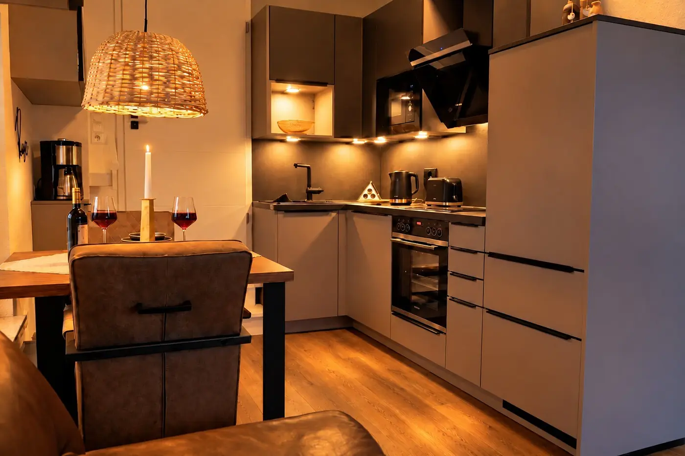 Küche und Essbereich der Ferienwohnung am Abend: Holztisch mit brennender Kerze und zwei Gläsern Rotwein, darüber eine Korbleuchte, dahinter die Küchenzeile mit Backofen und Kochfeld.