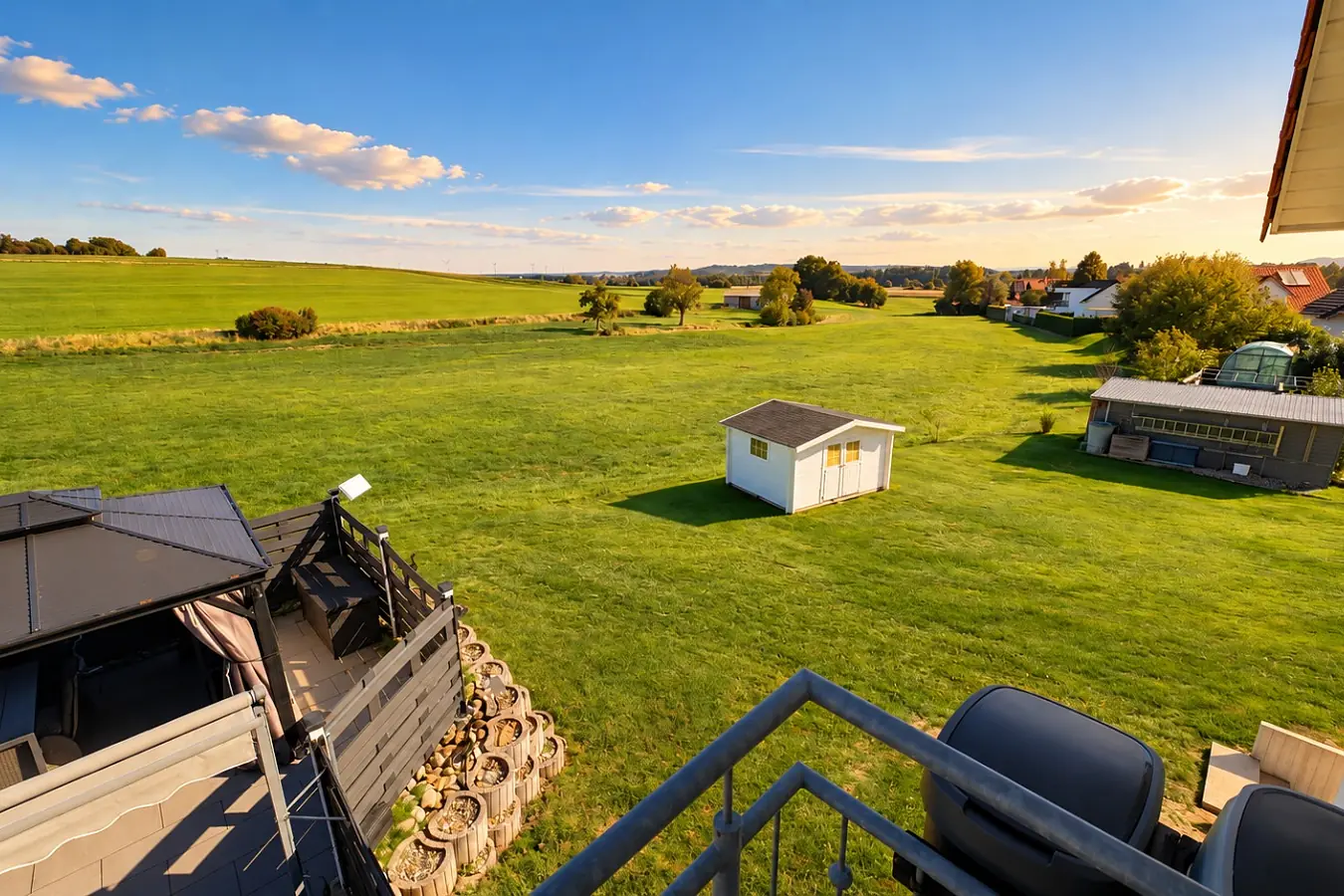 Blick von der Terrasse über Wiesen und Felder zum Ortsrand im Abendlicht, im Vordergrund das Geländer der Terrasse und ein kleines weißes Gartenhaus auf der Wiese.