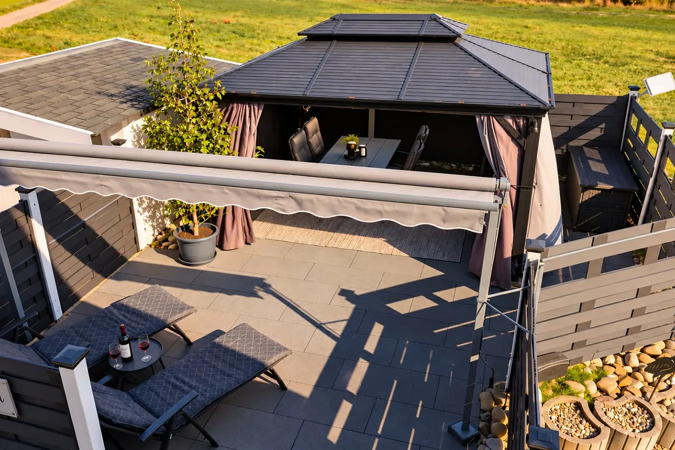 Die Terrasse der Ferienwohnung aus der Vogelperspektive: überdachter Pavillon mit Essplatz, ausgefahrene Markise, zwei Liegestühle mit Beistelltisch, ringsum ein dunkler Sichtschutzzaun, dahinter die Wiese.