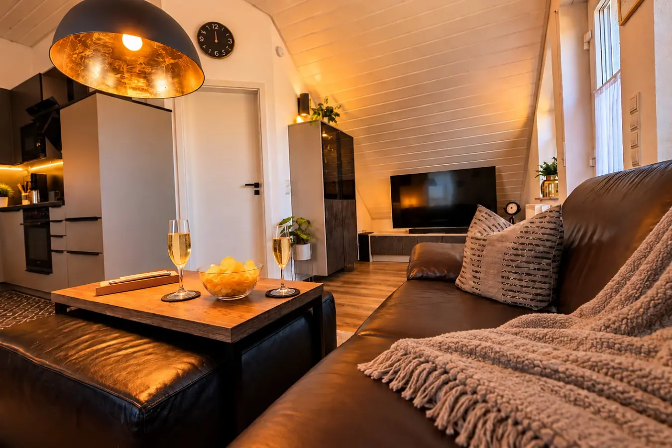 Der Wohnbereich der Ferienwohnung: braune Ledercouch mit Kissen und Wolldecke, davor ein Couchtisch mit zwei Gläsern Sekt und einer Schale Chips, gegenüber der Fernseher auf dem Sideboard, links die Küche.
