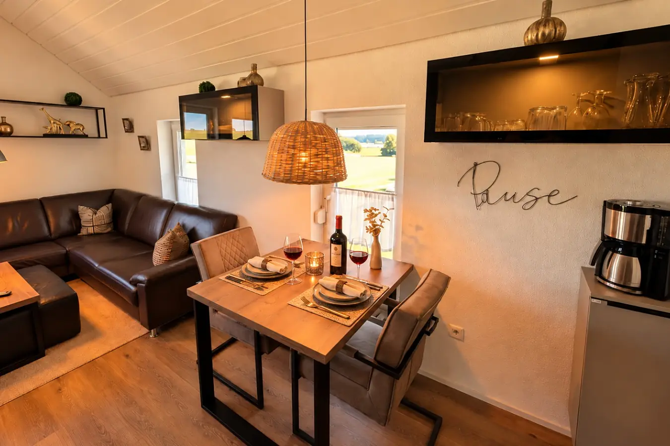 Wohn- und Essbereich von der Küche aus: gedeckter Holztisch mit Rotwein und Kerzen unter einer Korbleuchte, dahinter die Ledercouch, an der Wand der Schriftzug «Pause», rechts die Vitrine mit Gläsern.