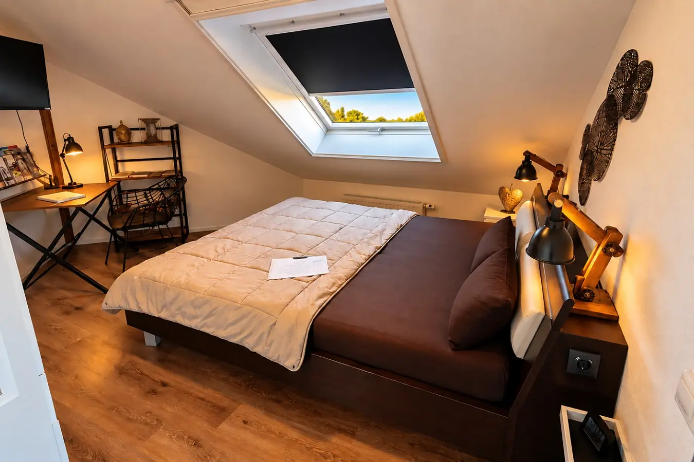 Schlafzimmer mit Doppelbett, dunkler Tagesdecke und zwei Leseleuchten am Kopfteil, links ein Schreibtisch mit Lampe und ein Metallregal, darüber ein Dachfenster mit Blick ins Grüne.