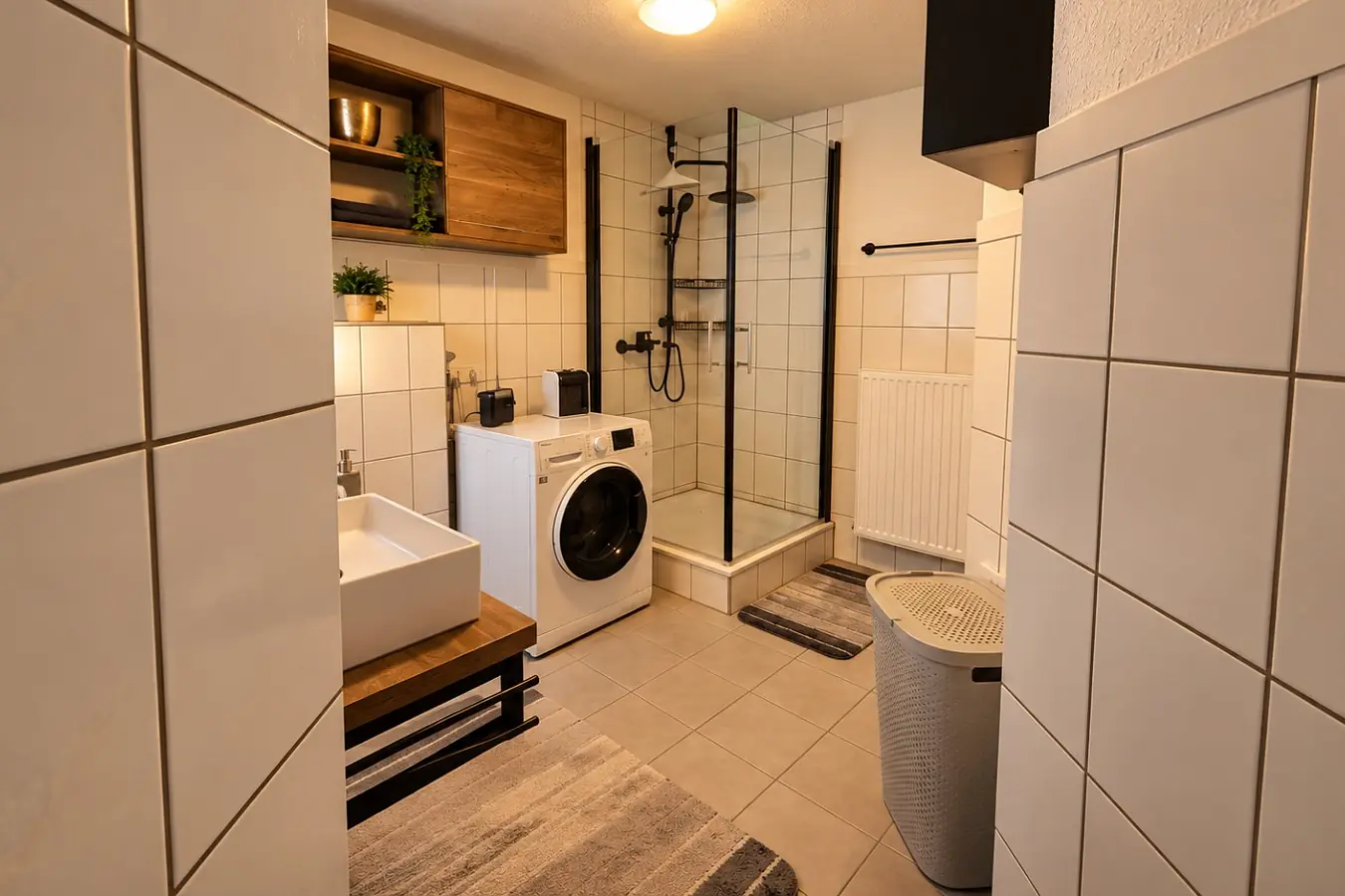 Bad der Ferienwohnung: Aufsatzwaschbecken auf einer Holzkonsole, daneben die Waschmaschine, dahinter die bodengleiche Dusche hinter Glas, rechts Heizkörper und Wäschekorb.