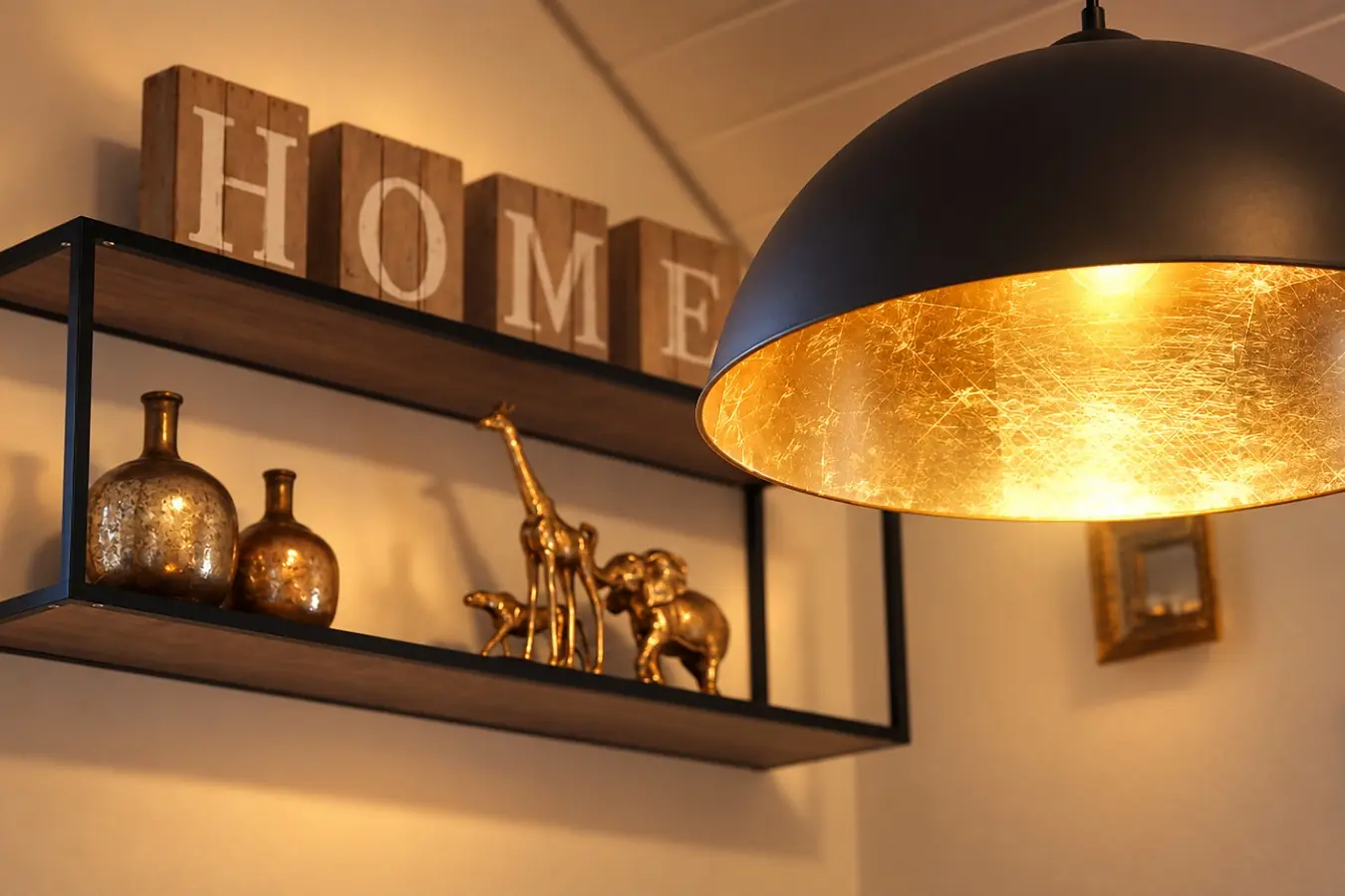 Schwarzes Wandregal mit goldener Deko und Holzbuchstaben «Home», daneben eine große schwarze Pendelleuchte mit goldener Innenseite.