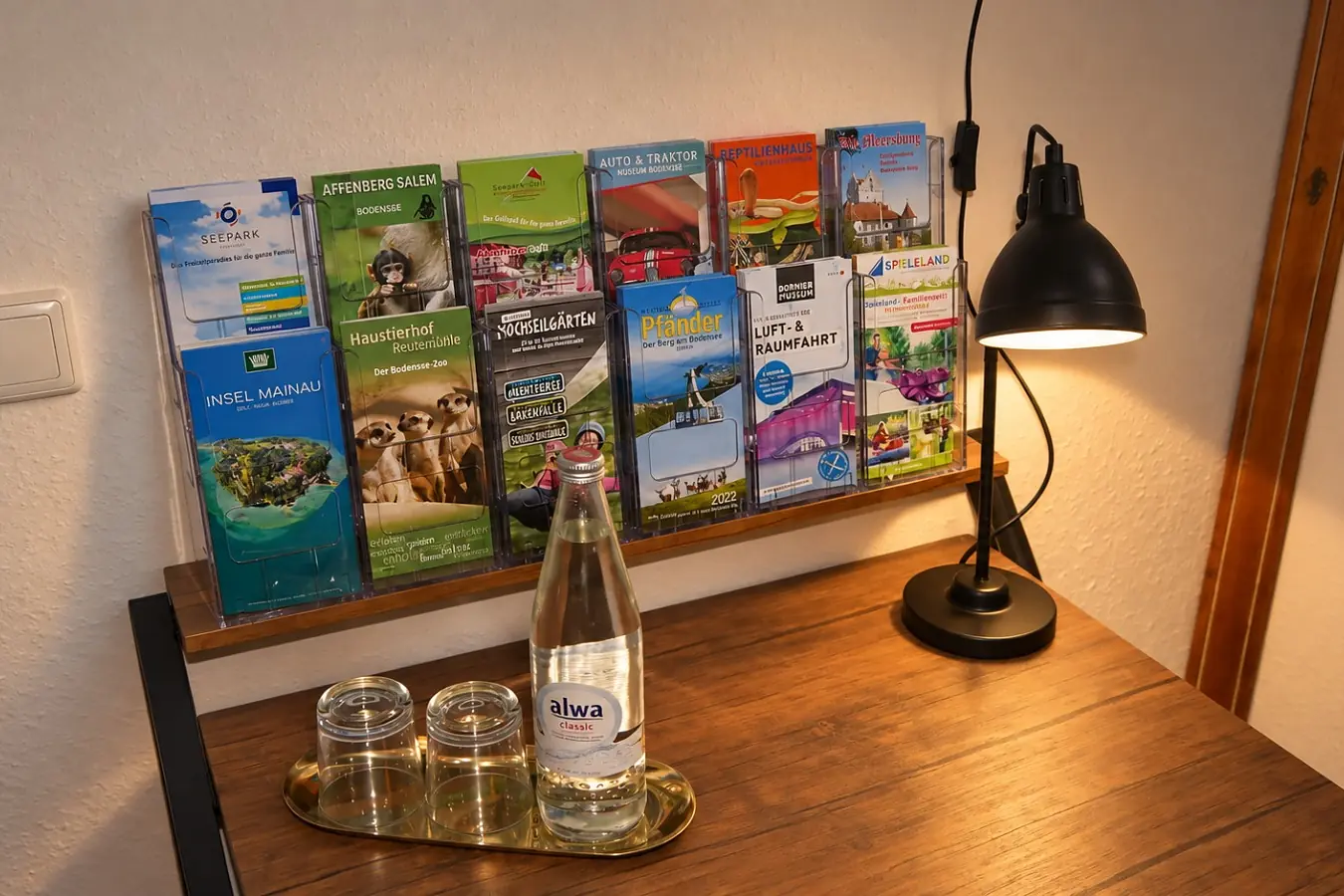 Prospektständer an der Wand mit Flyern zu Ausflugszielen in der Region, darunter ein Holztisch mit Leselampe, einer Flasche Wasser und zwei Gläsern auf einem goldenen Tablett.